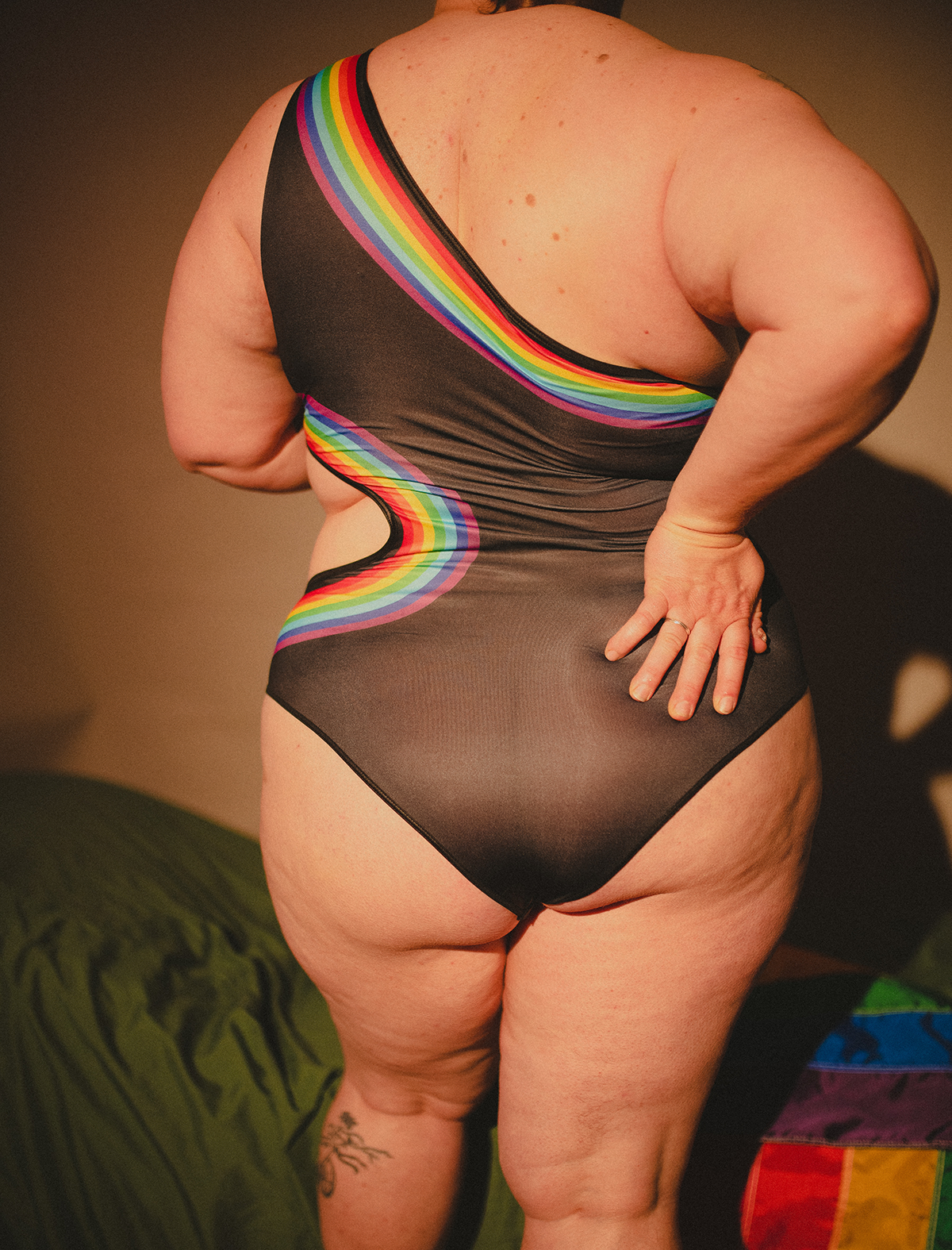 A queer person in a rainbow bodysuit turned away from the camera and showing off their ass
