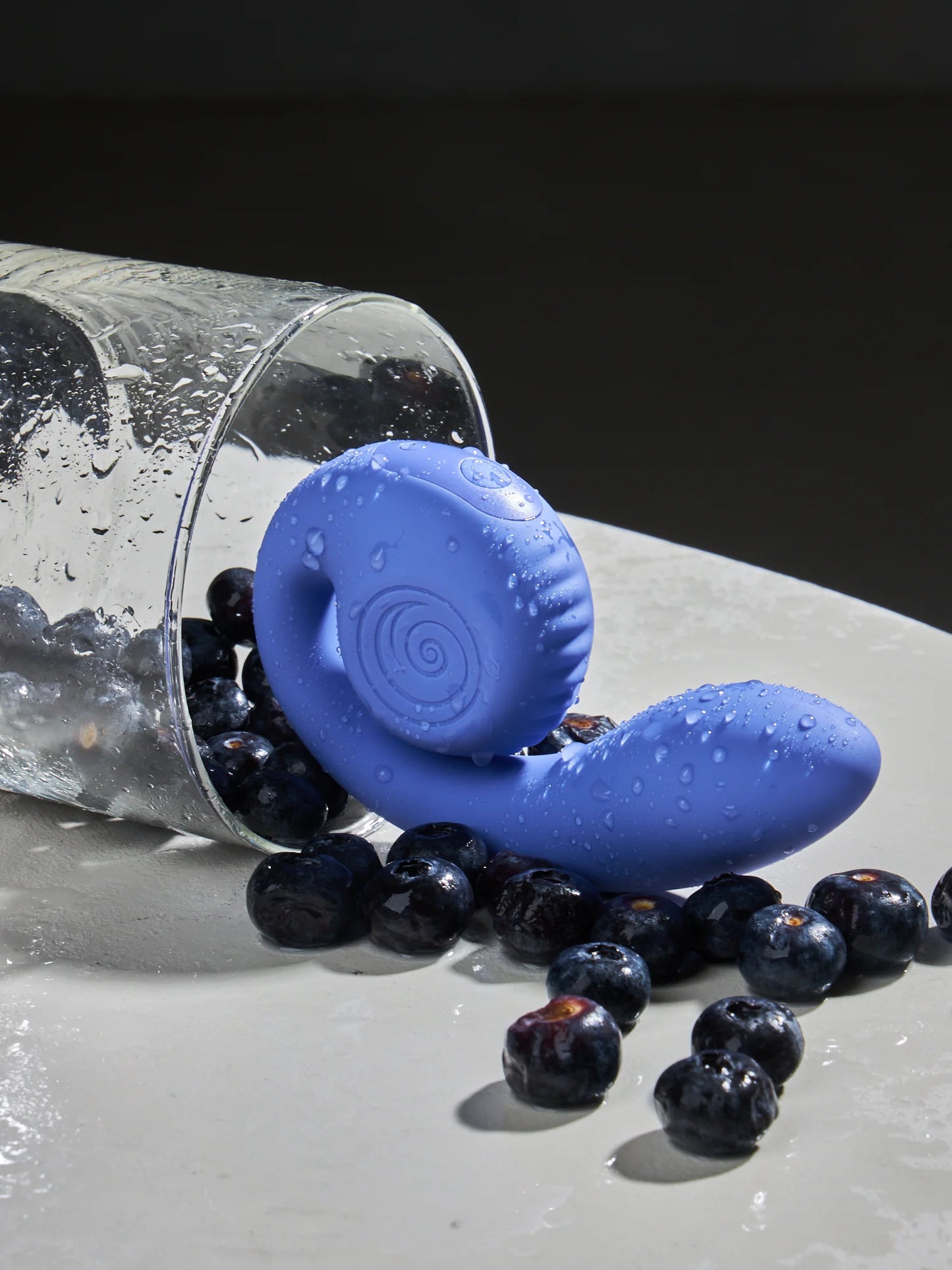 A blue vibrator with a snail-like design, placed next to a glass and scattered blueberries, indicating it's waterproof and splashproof.