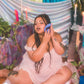Woman in a white dress sitting on the floor with a colorful curtain and plants in the background