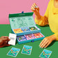 Person playing Biird x Jouissance Club Together Couples Card Game with colorful cards and a game box on a green table against a pink background