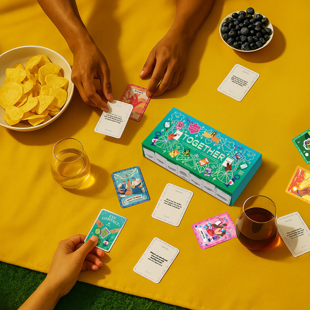 People playing Biird x Jouissance Club Together Couples Card Game with cards and snacks on a yellow tablecloth.