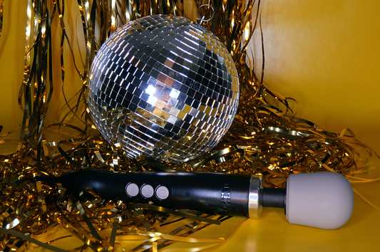 Doxy Original Massager in front of a disco ball and gold streamers. The Doxy Massager is a wand style vibrator that plugs into a wall with a thick grey head and a black body with large easy to use grey buttons