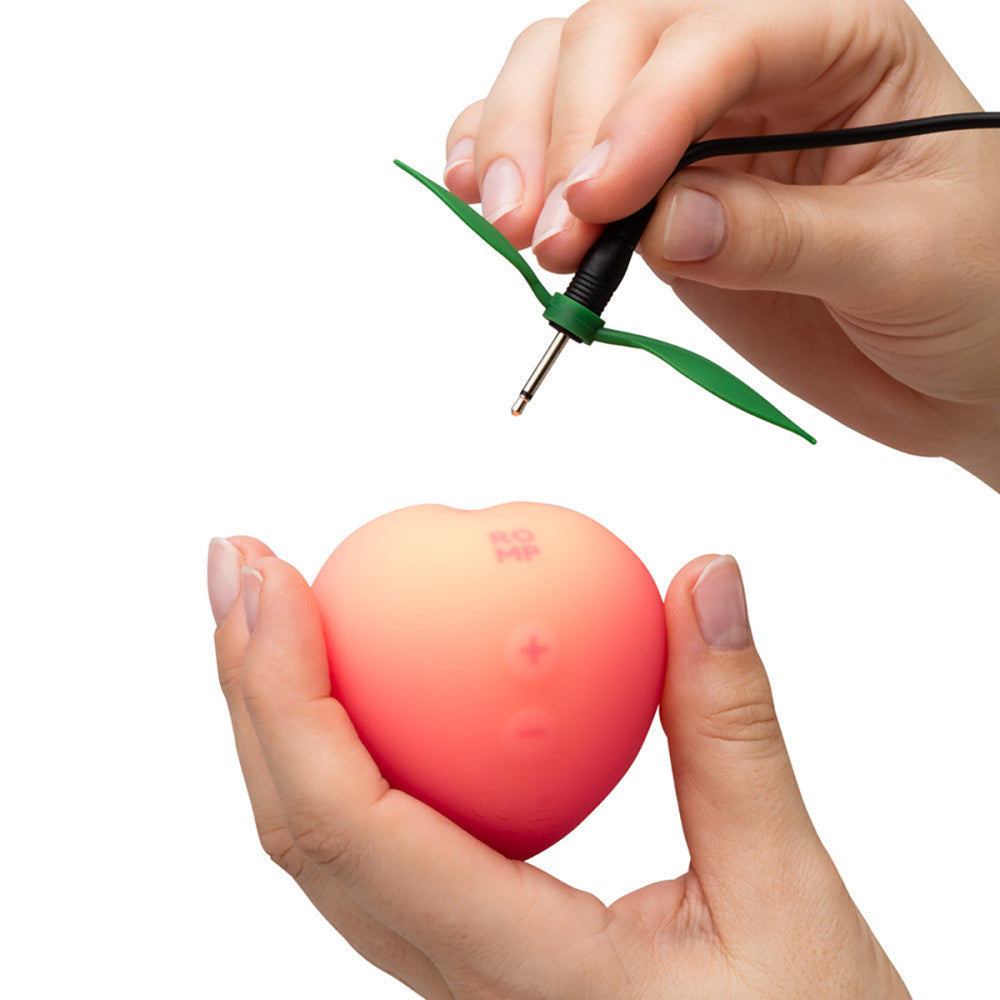 Romp Pink Peach Vibrator with a green and black cable being plugged into it, held by hands against a white background.