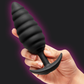 A hand holding a black ribbed silicone anal plug with a curved suction-cup base against a dark background with a purple glow effect.