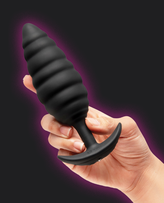 A hand holding a black ribbed silicone anal plug with a curved suction-cup base against a dark background with a purple glow effect.