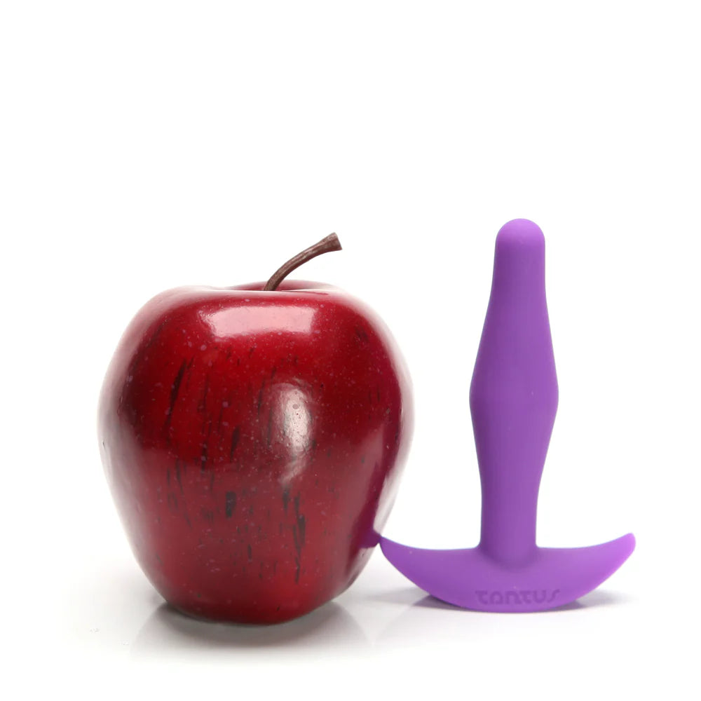Red apple next to a purple silicone Tantus Little Flirt Anal Plug on a white background