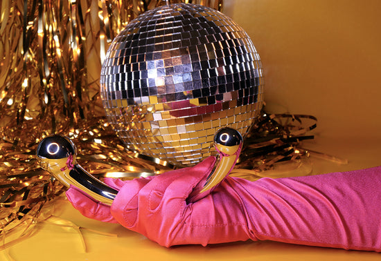 Njoy Pure Wand is a stainless steel curved dildo with two doifferent sized bulbs at the ends for g-splot pleasure. This toy is being held by a pink gloved hand in front of a discoball and gold streamers. 