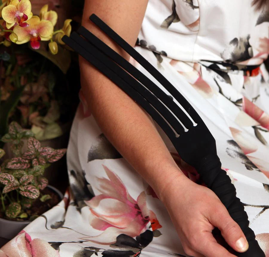 Person holding a black Tantus Tawse Paddle with floral-patterned clothing and plants in the background