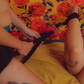 A person is using the Sportsheets Under The Bed Restraint System to restrain another person who is lying on a bed with a colorful floral background. The restraint system includes black straps and cuffs.